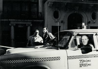 Announcements and invitations depicts: A man driving in a taxi looks out to the camera, two men stand behind looking to the camera as well