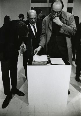 Photographic Materials depicts: Three men (one of them Milton Glaser) looking at the contents of a black binder on a white plinth