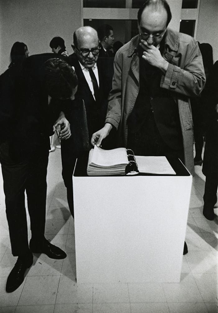 Photographic Materials depicts: Three men (one of them Milton Glaser) looking at the contents of a black binder on a white plinth