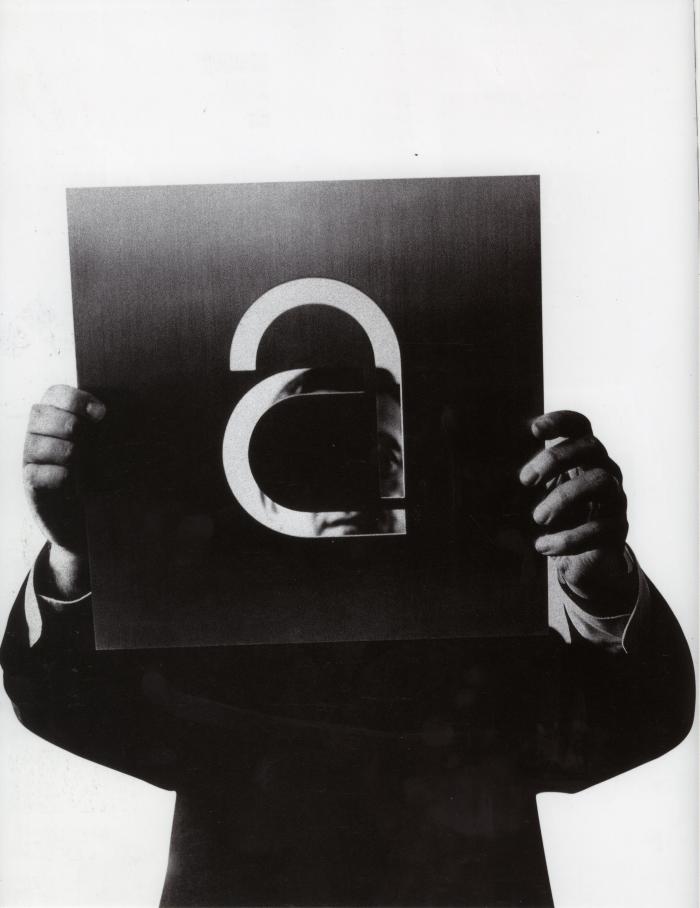 Photographic Materials, Logos and identities depicts: A man holds a piece of metal with a large lower case "A" carved out of it in front of his face, you can see parts of his face through the openings in the metal 