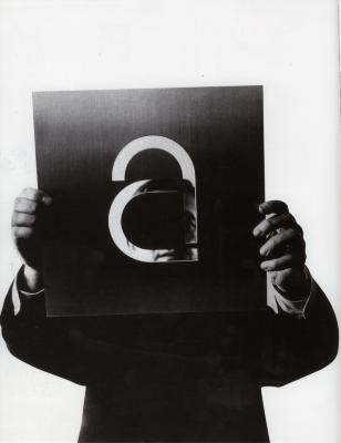 Photographic Materials, Logos and identities depicts: A man holds a piece of metal with a large lower case "A" carved out of it in front of his face, you can see parts of his face through the openings in the metal 