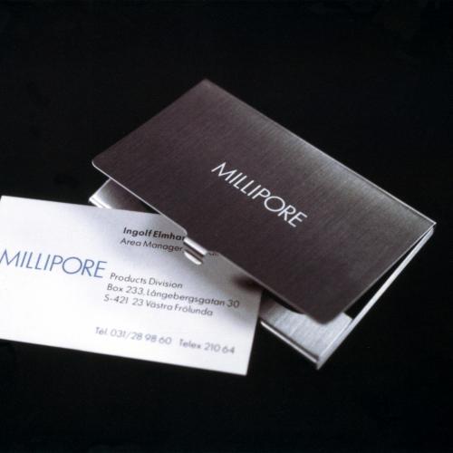 Three-Dimensional Objects, Logos and identities depicts: A metal business card holder with "Millipore" engraved on the front, a Millipore business card sticks out of it 