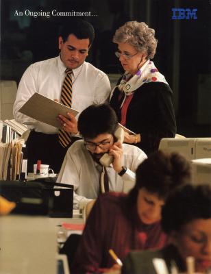 Two people look at a clipboard, in front of them sits a man talking on the phone 
