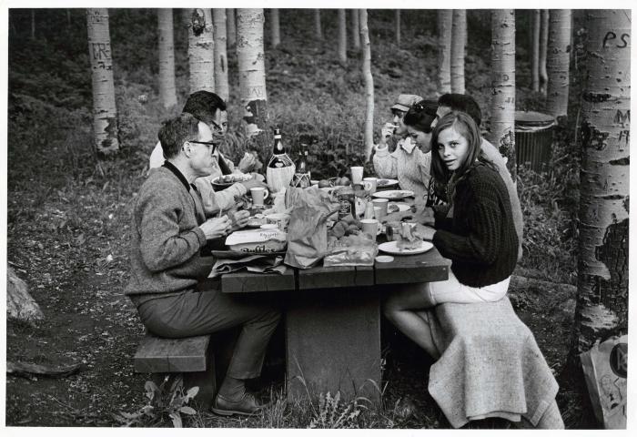 Photographic Materials depicts: Black and white photo of a group at a picnic table in the woods with an elegant spread. A young woman at the table looks directly at the camera