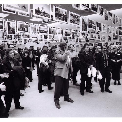 Black and white photo of large, somber crowd at here is NY exhibition, many photos are hung from clips around them. A man in the center has his hand over his mouth.  