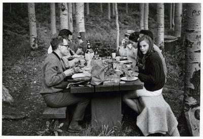 Photographic Materials depicts: Black and white photo of a group at a picnic table in the woods with an elegant spread. A young woman at the table looks directly at the camera