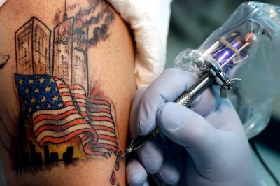 Photographic Materials depicts: closeup of a hand holding a tattoo gun, tattooing an arm with a tattoo of the twin towers, one in flames, and a torn american flag