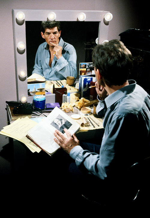 Photo of Ian McKellen behind the stage, practicing in front of a messy desk and mirror.