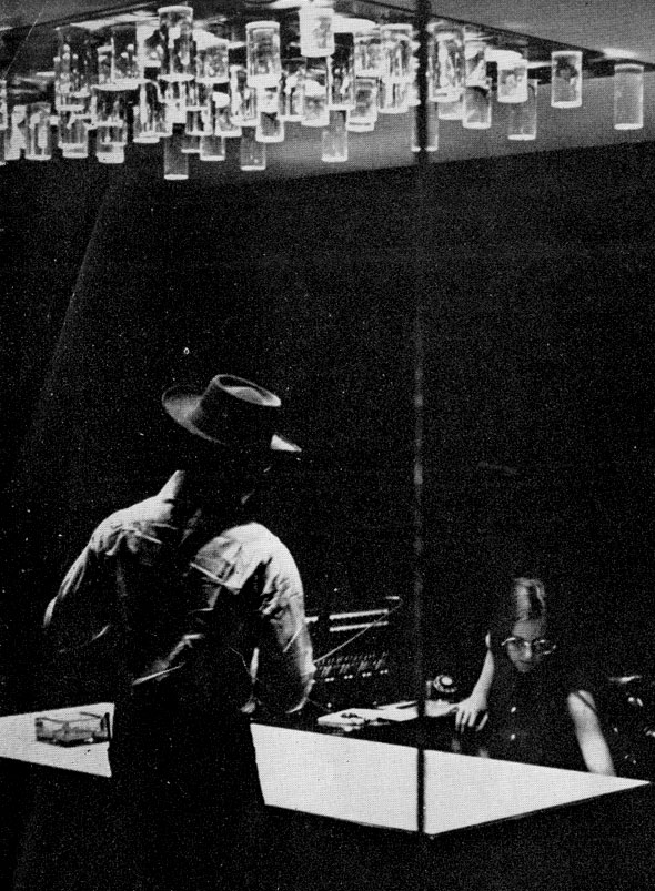 Black and white photograph of mid-century agency; a man in a hat approaches a woman at her desk underneath an expressionistic light fixture in the reception room.