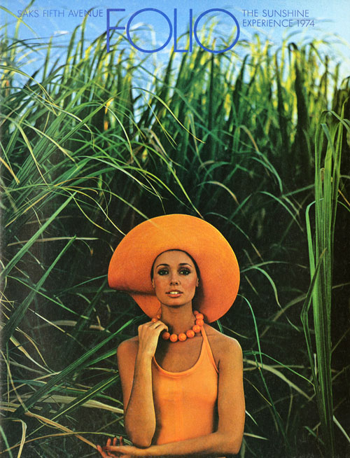 Bright color photo of a woman wearing all orange wide-brim hat and beads standing against tall green plants