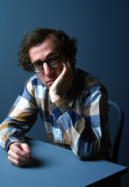 Photo Portrait of Woody Allen, seated in a chair and leaning against a vacant desk.