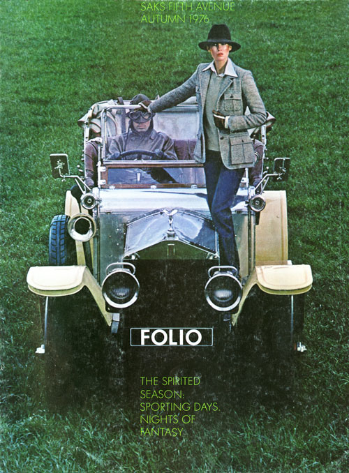 Color photo of a woman in a hat and blazer standing on top of a 1930's convertible car being driven by a man with goggles in a green field.