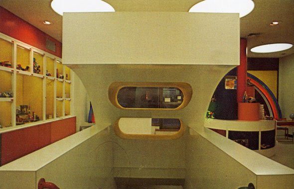 A photo of a a white geometric-like staircase housing inside the location. Its behind the front desk. It has a red railing on the left and a blue railing on the right. The house has a curved top that has two wide holes that serves as shelfs for the front desk. You see the cubbies of toys on the left with rainbow striped colored walls. 