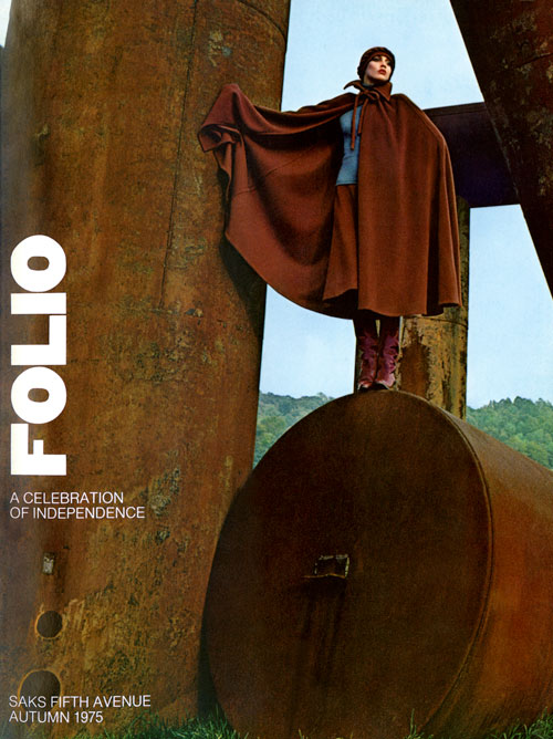 Color photo of an elegant woman in a long brown cloak standing atop some rusted metal structures in a field