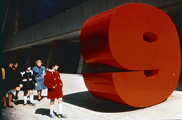 Five people looking at a large red sculpture of the number nine.