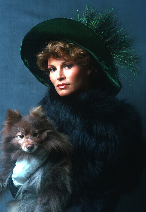 Photo Portrait of Racquel Welch wearing a green floppy hat, faux fur, and holding a brown Pomeranian dog.