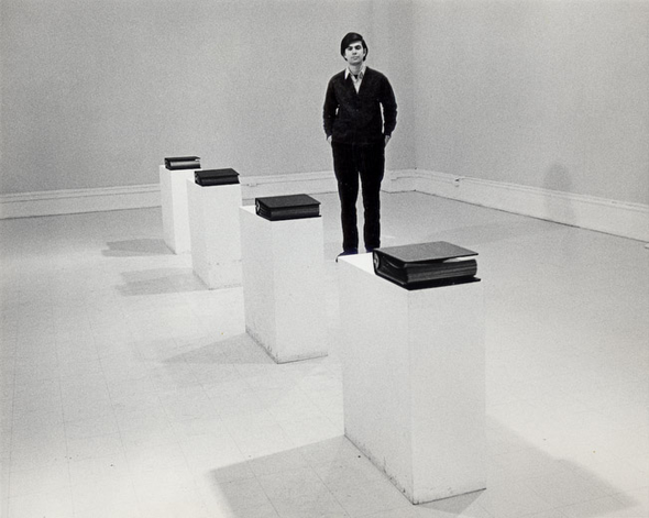 Black and white photo of a man standing in an art gallery; there are 4 binders mounted on pedestals